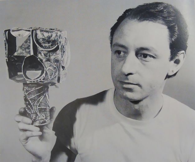 Black and white photograph. Shown from the chest up, aa pale-skinned man with his dark hair brushed back from his forehead and wearing a white t-shirt holds a sculpture in one hand. He looks to our left with dark eyes past the sculpture, which is a cube on top of a cylinder. The abstracted head, the cube, has exaggerated eyes and a wide-open mouth. The entire work is collaged.
