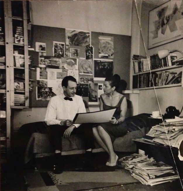 Black and white photograph. Two pale-skinned people sit on a bed and hold a piece of paper about the size of a baking sheet together. The man wears a white button-down and black tie. The woman a sleeveless dress and heels. Papers are piled on the floor and shelves around them, and images are pinned on a board behind.