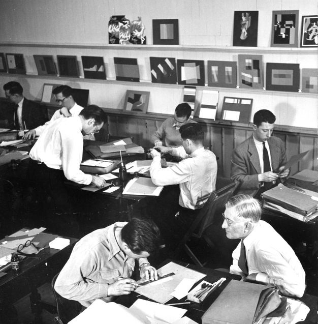 Black and white photograph. We look down onto seven young, clean-shaven men working with paper at tables. An eighth, with glasses and silvery hair, watches the man closest to us.